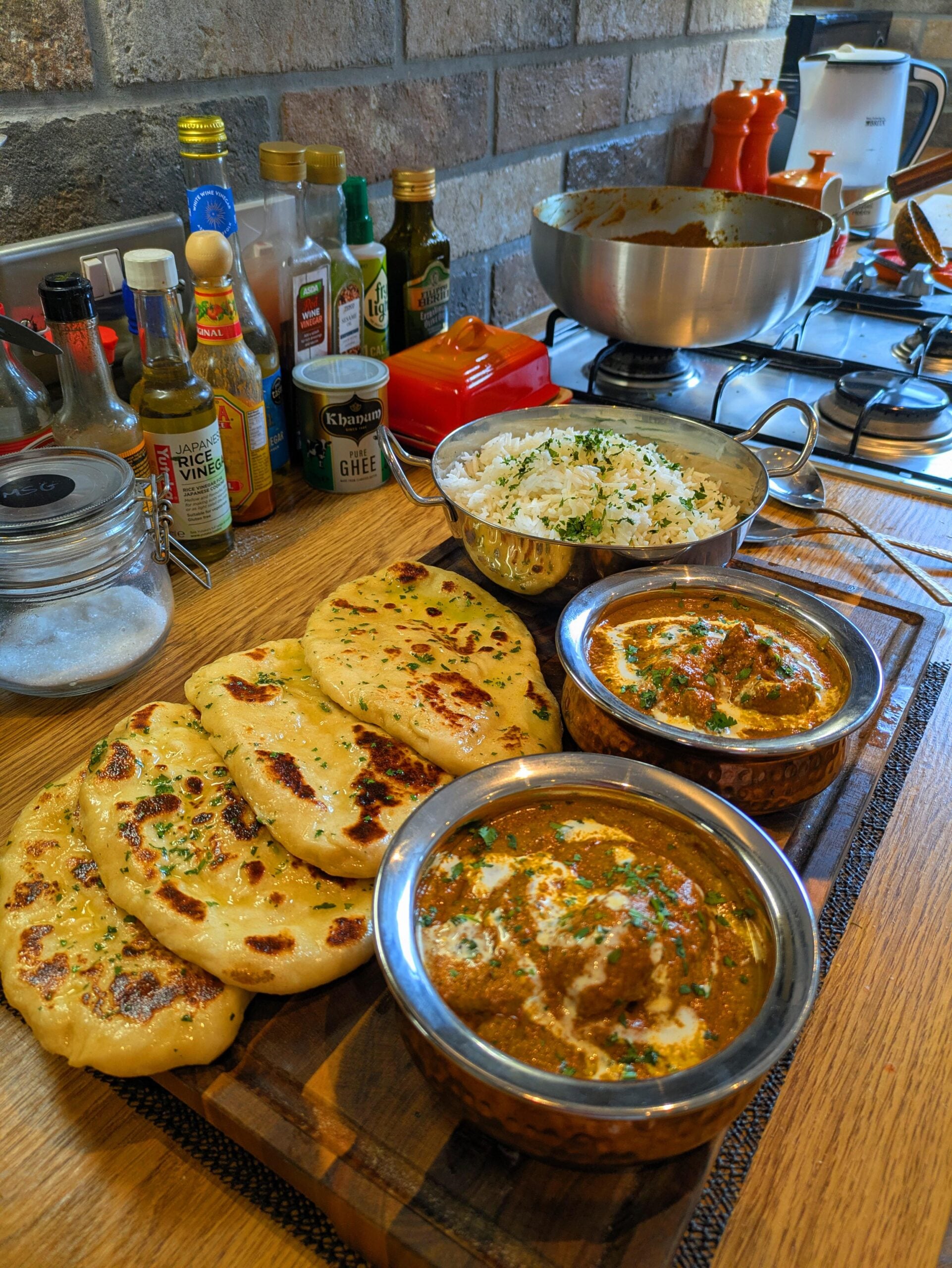 A wooden board with naan bread, two bowls of curry, and a bowl of rice on a kitchen counter with spices, oil bottles, and a large metal mixing bowl in the background.