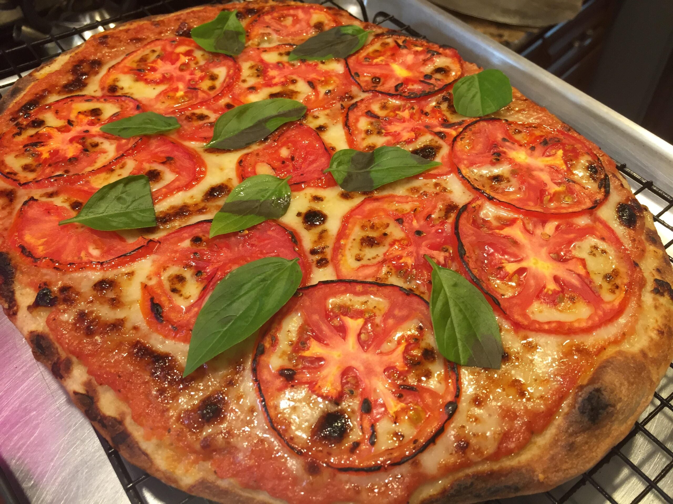 A freshly baked pizza topped with melted cheese, sliced tomatoes, and fresh basil leaves rests on a cooling rack.