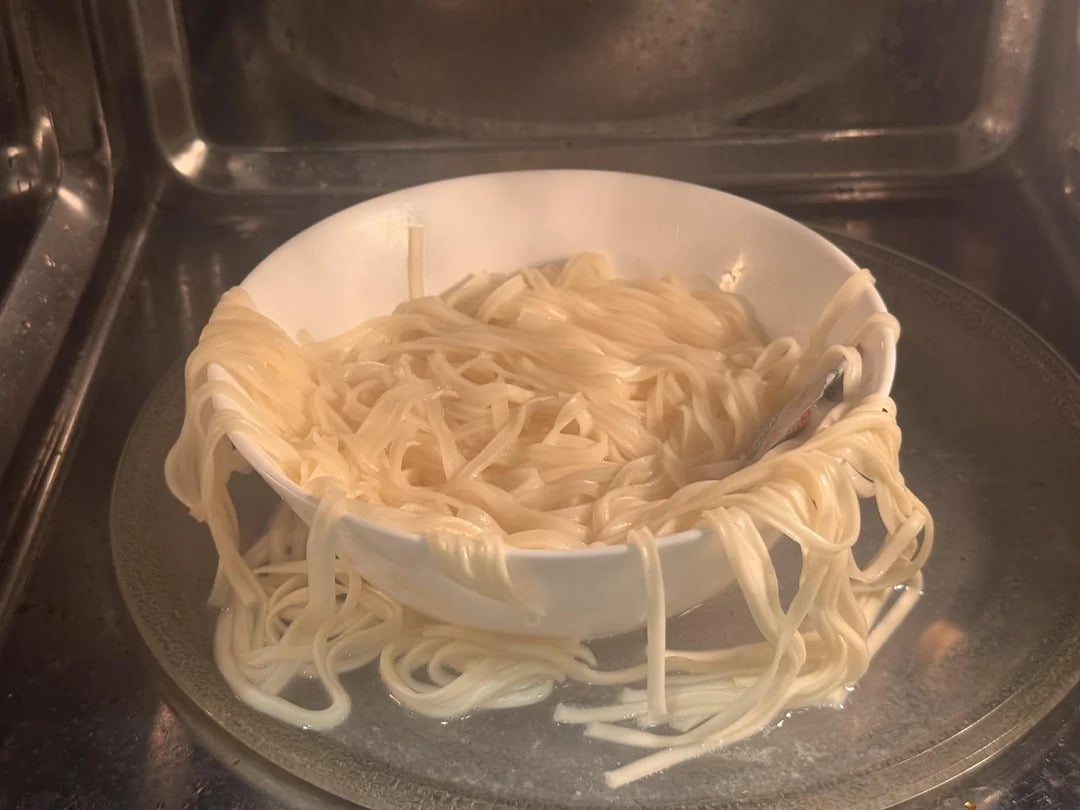 A bowl of noodles in a microwave has overflowed, with cooked noodles spilling over the rim and onto the microwave plate.