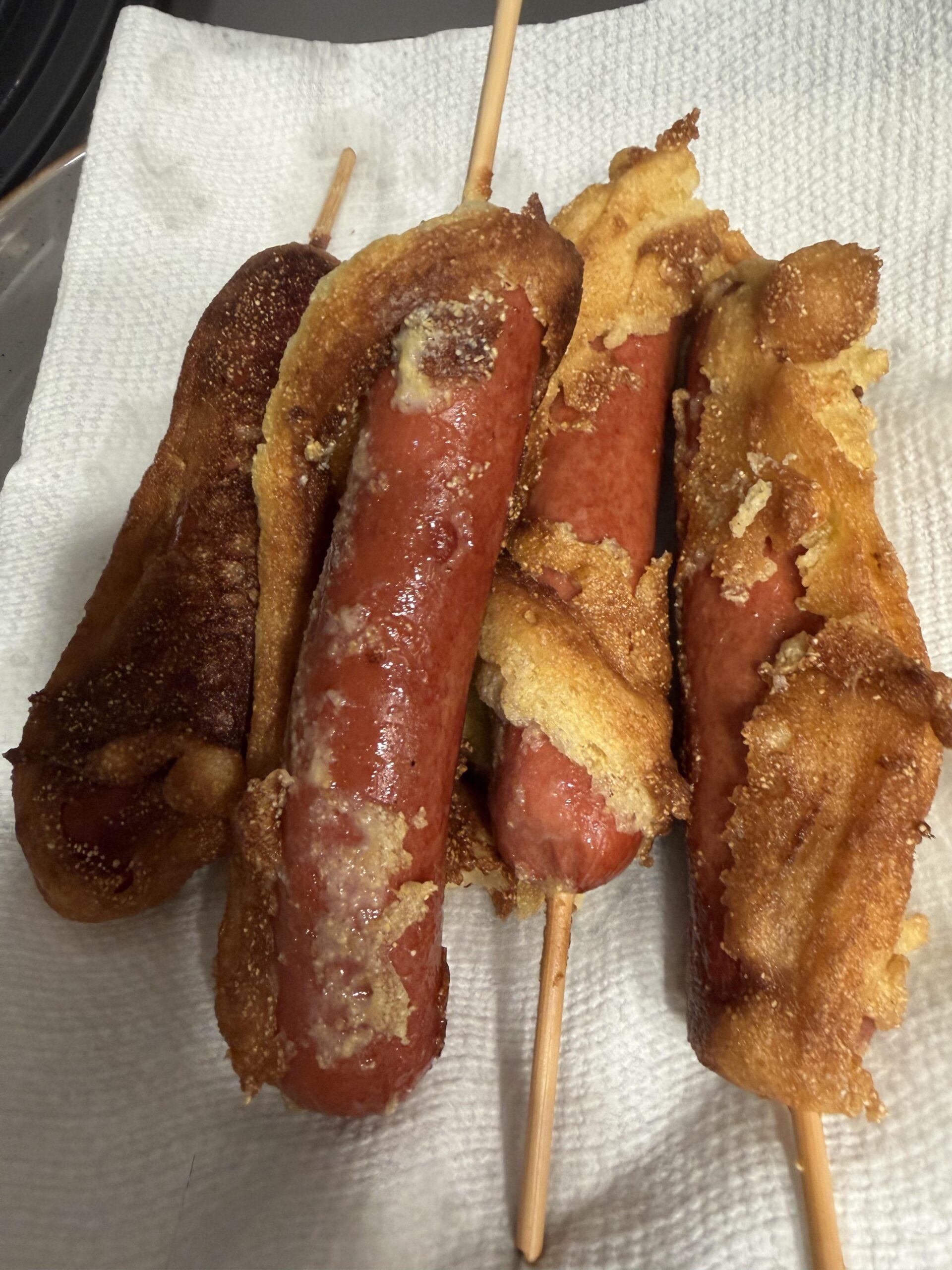 Four corn dogs with uneven, golden-brown batter rest on a white paper towel. The batter appears homemade and crispy in some areas, with frayed edges and visible texture.