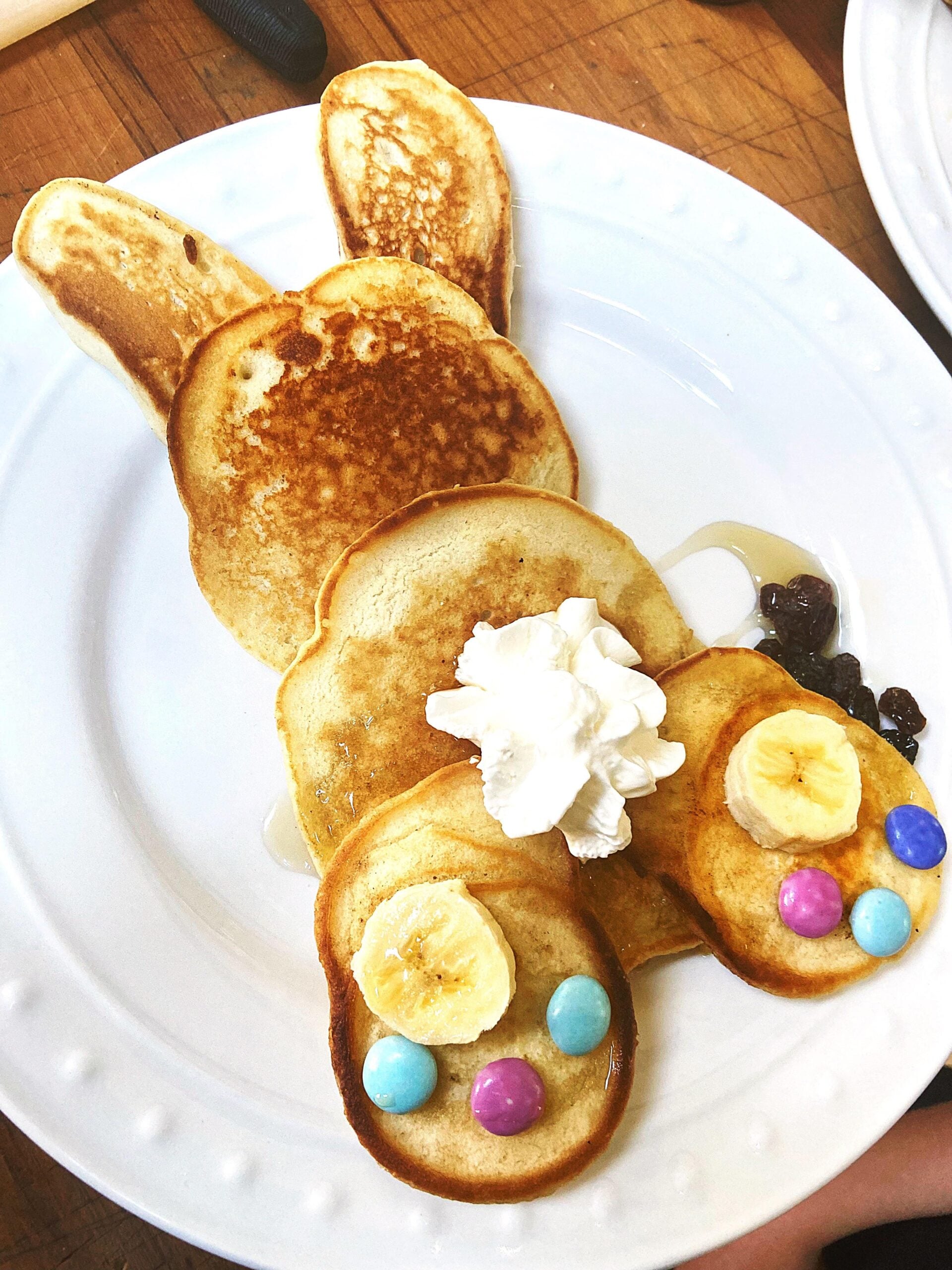 A bunny-shaped homemade pancake with banana slices, colorful candies, whipped cream, and syrup on a white plate.