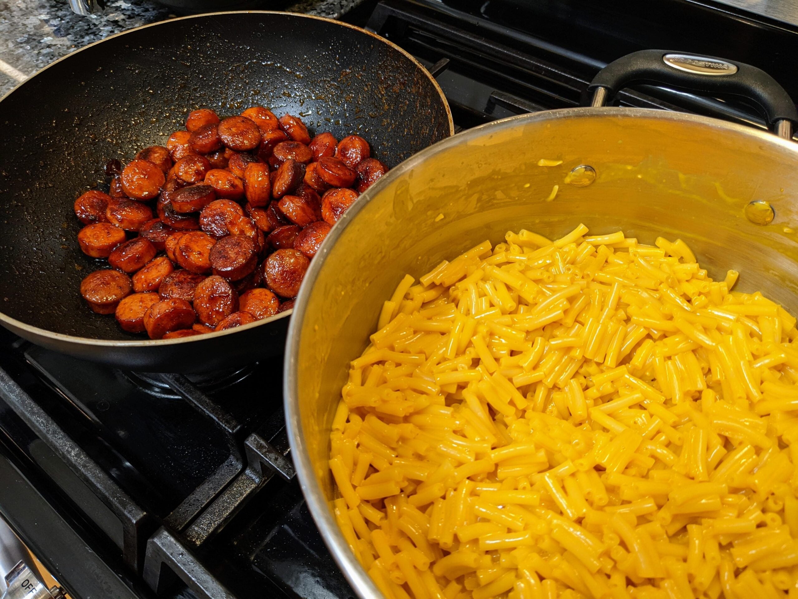 A pot of cooked macaroni and cheese next to a skillet filled with sliced, browned sausage on a stovetop.