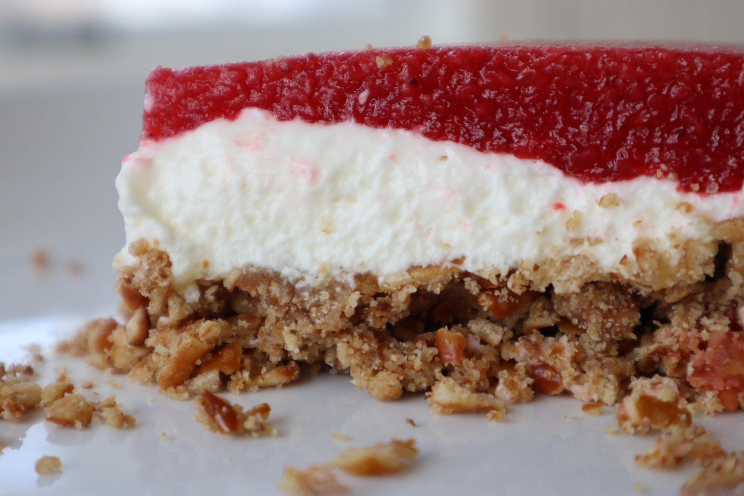 A close-up of a homemade dessert with a crumbly nut base, a thick layer of creamy white filling, and a smooth red fruit topping on a white plate.