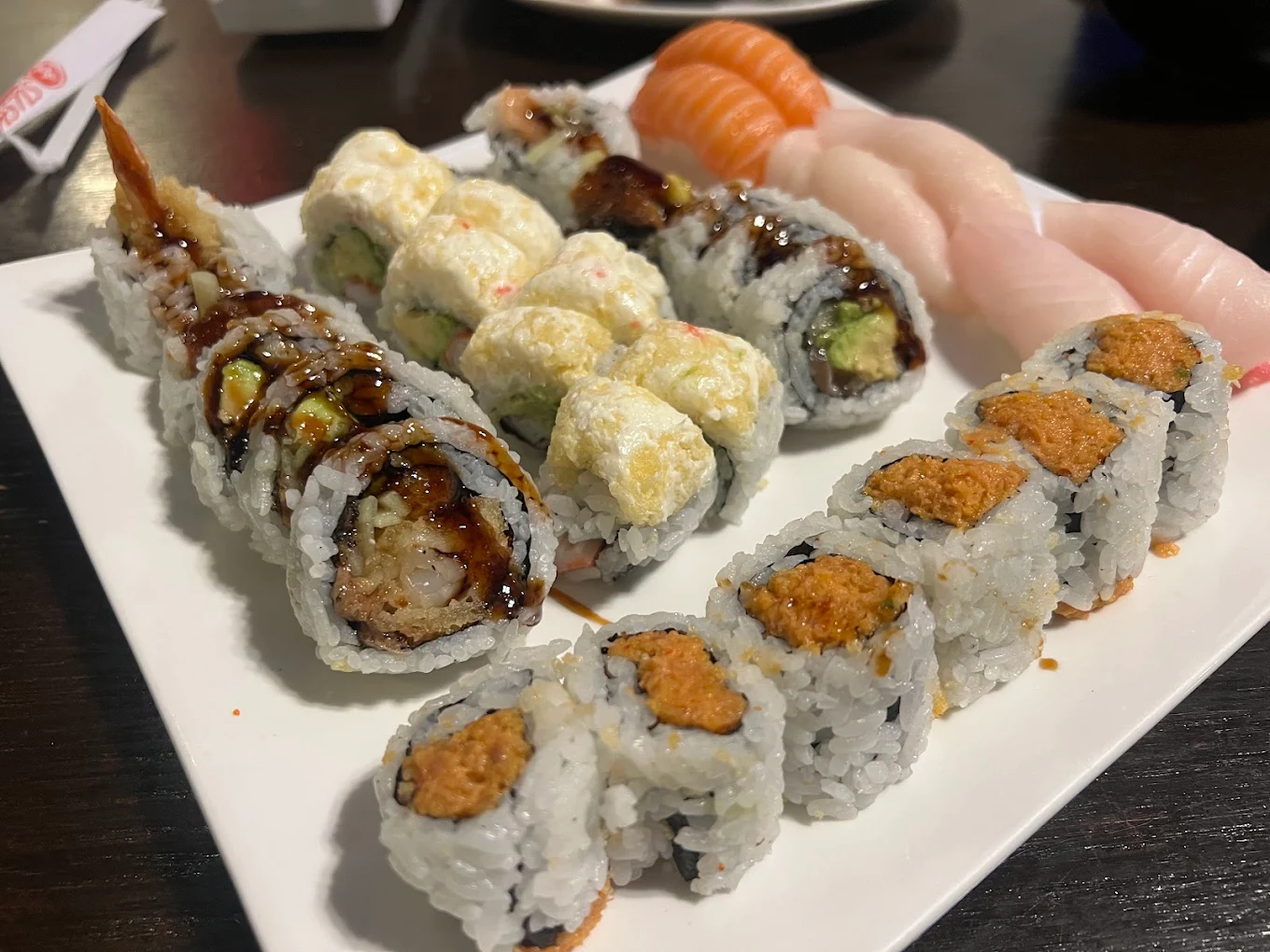 A white plate with assorted sushi rolls and nigiri, including shrimp tempura rolls, sushi topped with sauce, pieces with avocado, and slices of salmon and white fish over rice. Chopsticks are visible in the background.