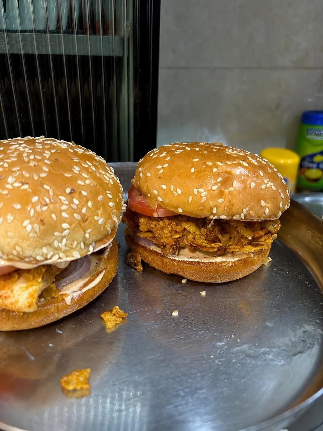 Two chicken burgers with sesame seed buns sit on a metal tray. The burger in front has lettuce, tomato, and crispy fried chicken. Condiments are visible in the background.
