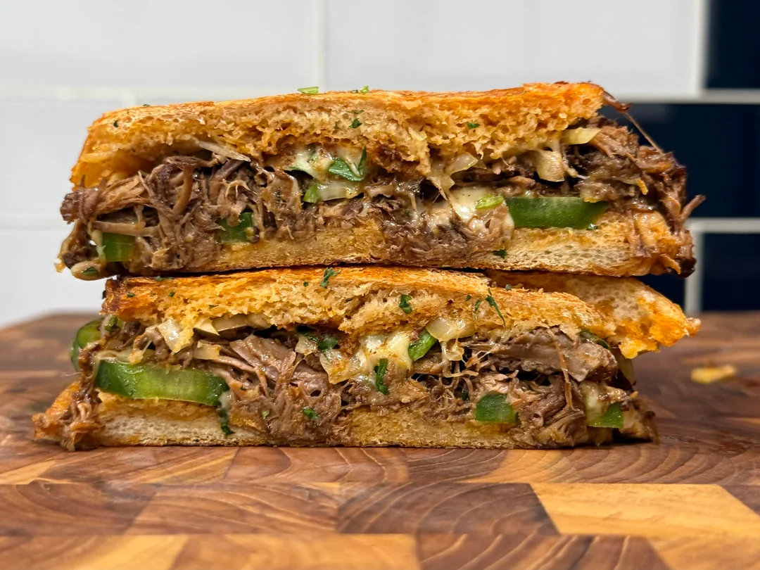 A close-up of a stacked sandwich with shredded beef, green peppers, onions, and melted cheese on toasted bread, placed on a wooden surface.