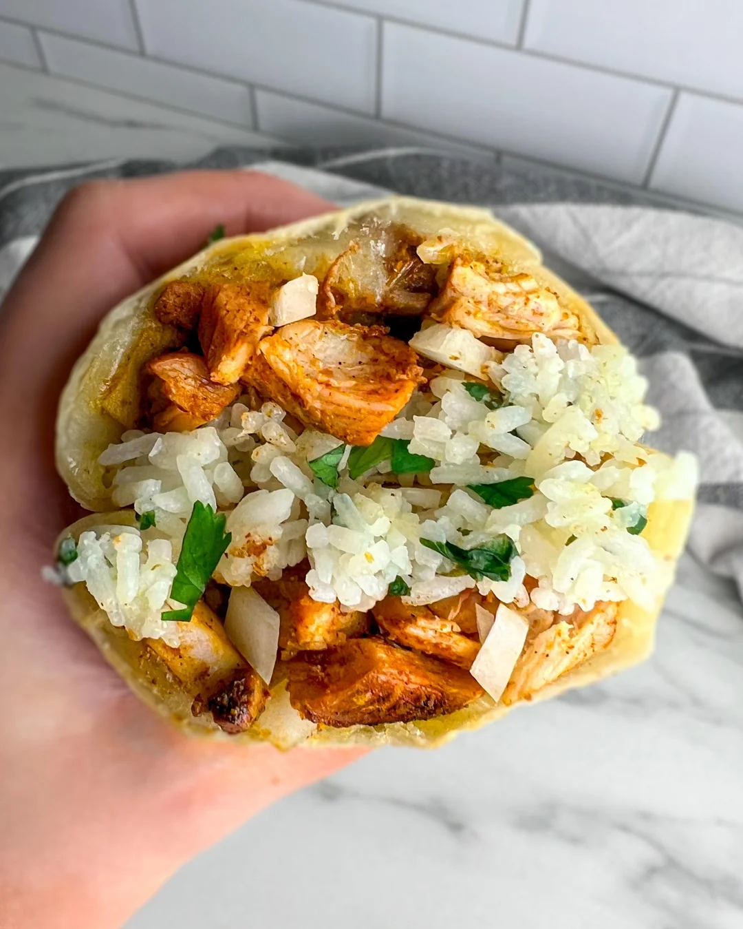 A hand holds a burrito filled with white rice, seasoned chicken, onions, and fresh cilantro against a marble surface and tiled wall background.