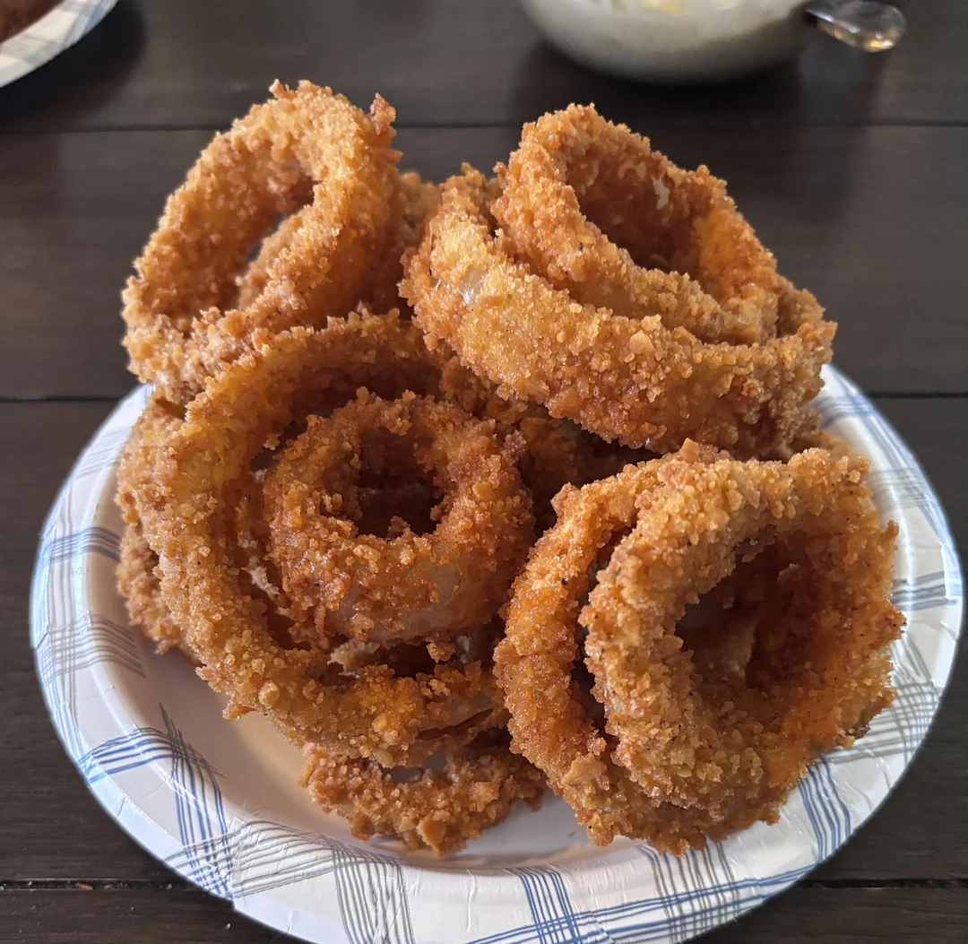 A stack of crispy, golden-brown onion rings is arranged on a white paper plate with blue patterns, set on a dark wooden table.