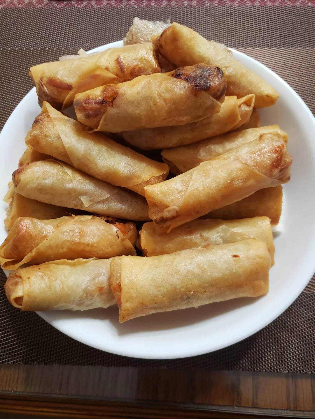 A white plate filled with neatly arranged golden-brown spring rolls, placed on a brown woven placemat.