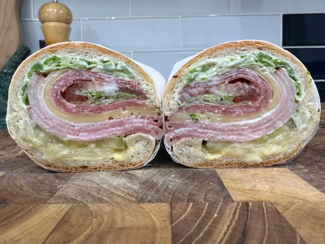 A cross-section of a sandwich with layers of lettuce, mayonnaise, sliced meats, cheese, and bread, displayed on a wooden cutting board in a kitchen setting.