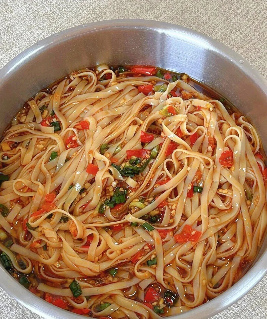 A bowl of flat rice noodles in a spicy soy-based sauce, mixed with chopped red chilies and green onions, sits on a light textured surface.
