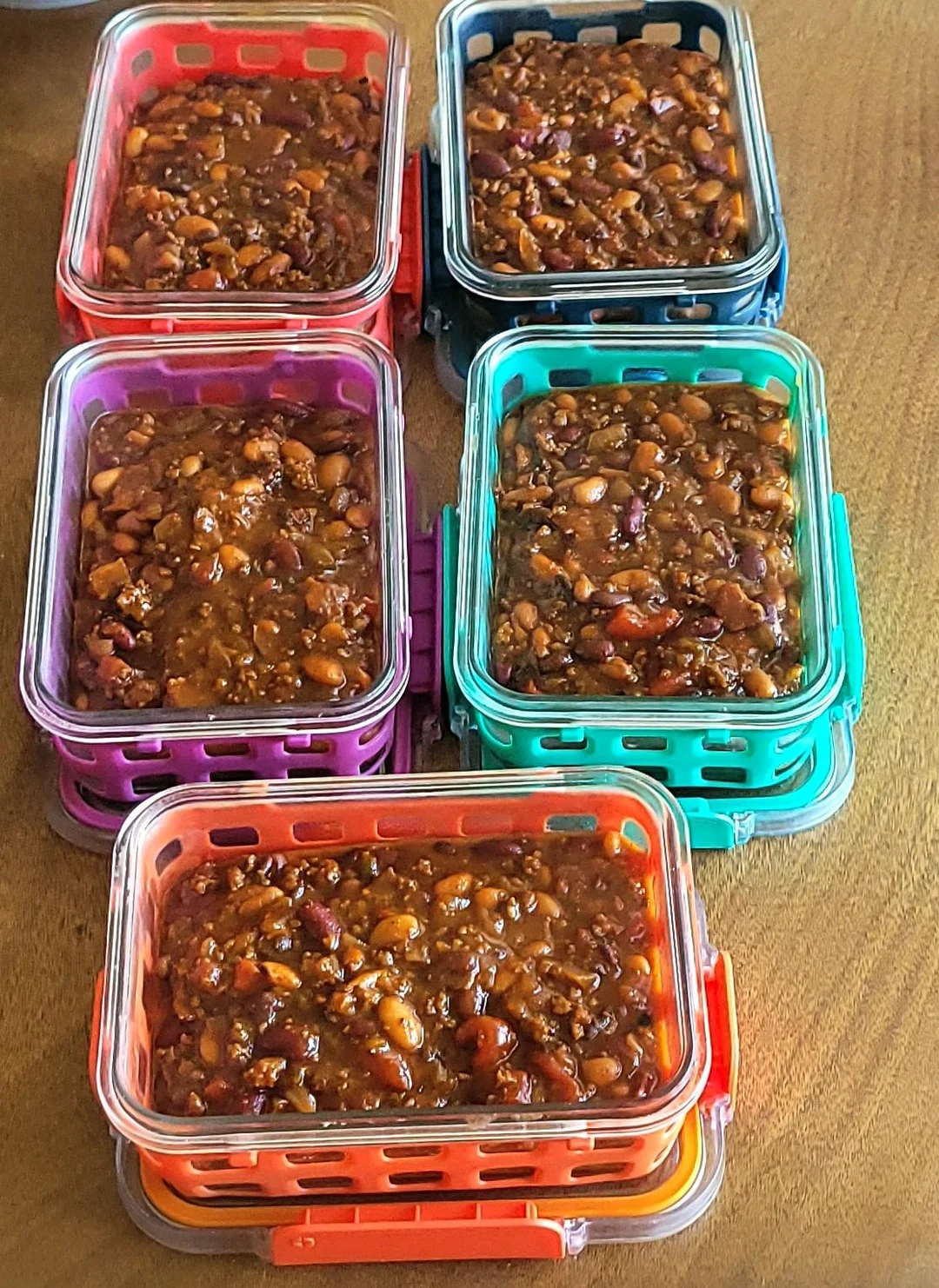 Five rectangular containers filled with mixed beans in sauce are arranged on a wooden surface. Each container has a different colored rim: purple, blue, red, green, and orange.