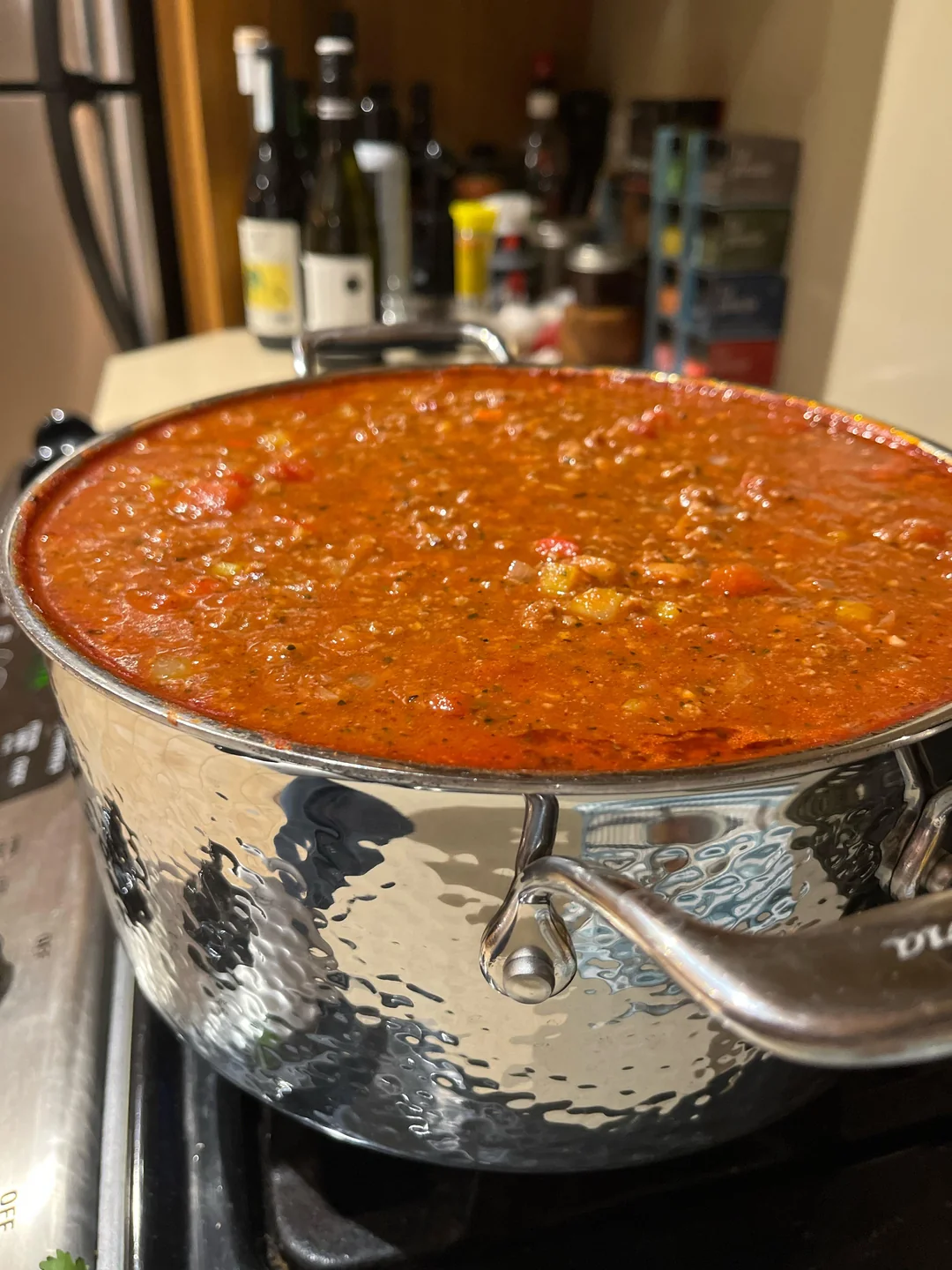 Large stainless steel pot filled to the brim with thick, chunky red tomato sauce on a stovetop; bottles and kitchen items are visible in the background.