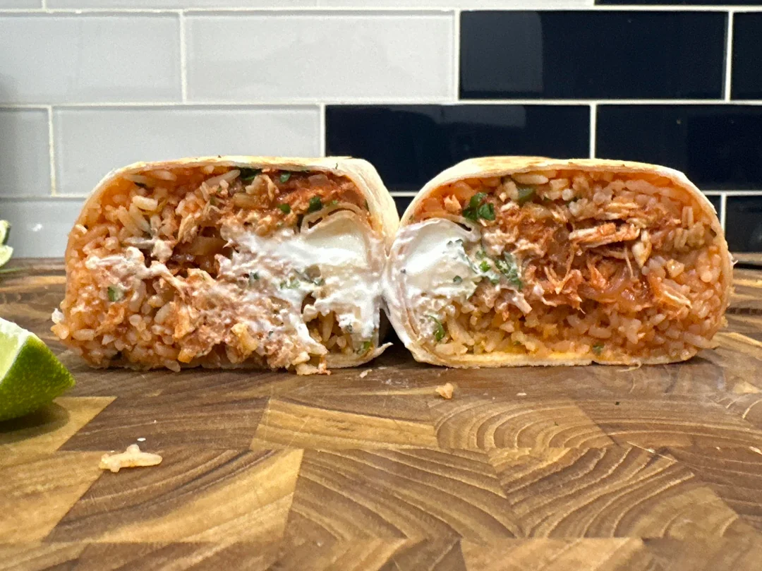 A burrito cut in half on a wooden surface, showing rice, shredded meat, cilantro, and sour cream inside, with tile backsplash and lime wedges in the background.