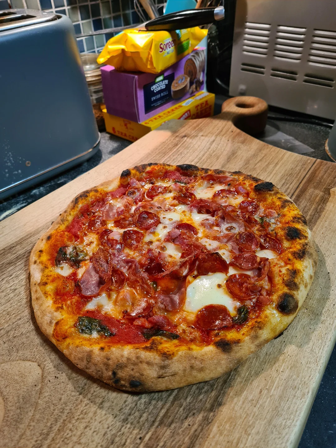 Homemade meals. A freshly baked pepperoni pizza with melted cheese sits on a wooden cutting board in a kitchen, with snacks and appliances visible in the background.