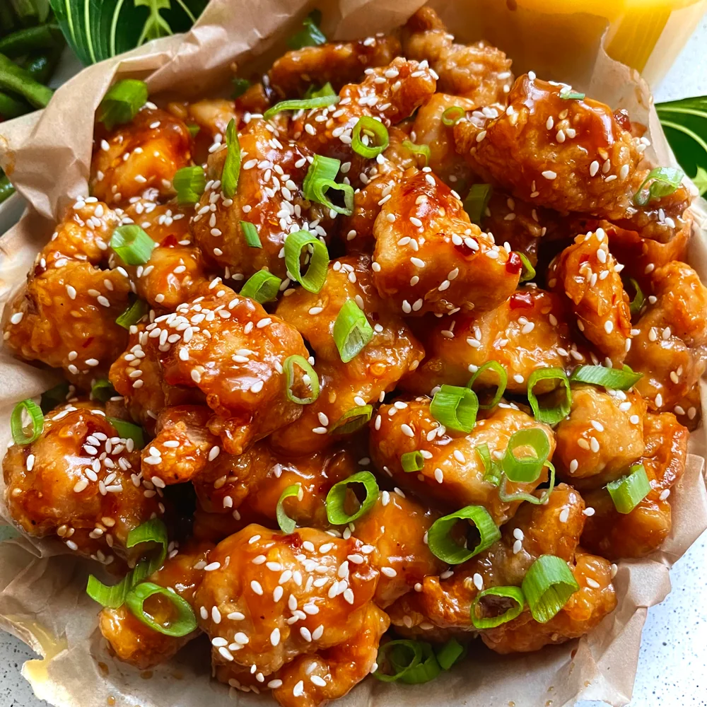 A basket of crispy orange chicken pieces glazed in sauce, garnished with chopped green onions and sesame seeds, served on parchment paper.