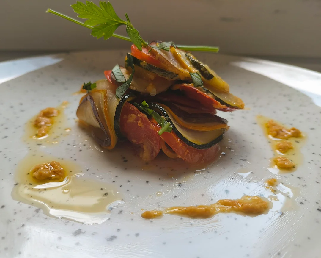 A small stack of thinly sliced vegetables, garnished with herbs and surrounded by drizzles of sauce, is artfully arranged on a white, speckled plate.
