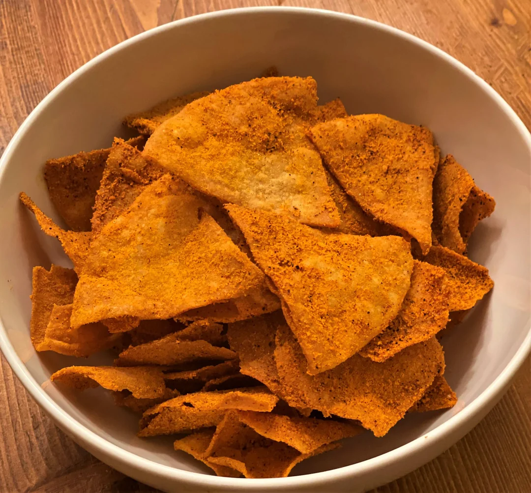 A white bowl filled with crispy, orange tortilla chips seasoned with a spicy red powder sits on a wooden table.
