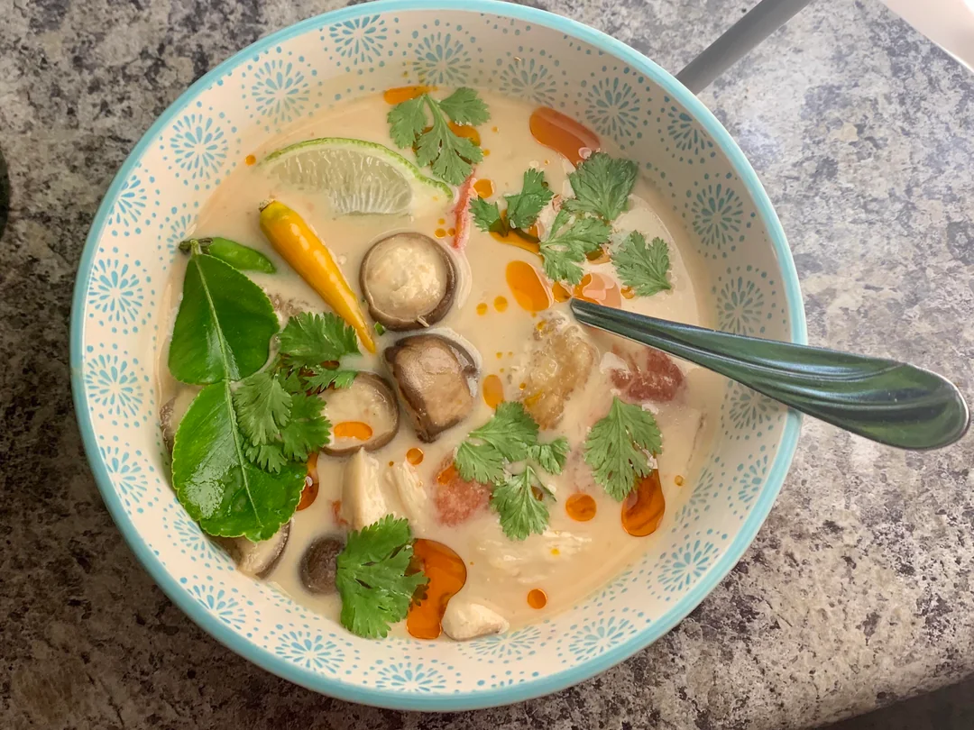 A bowl of Thai soup with mushrooms, lime, cilantro, chili peppers, kaffir lime leaves, and red oil drops in a creamy broth, with a spoon in the bowl on a granite countertop.