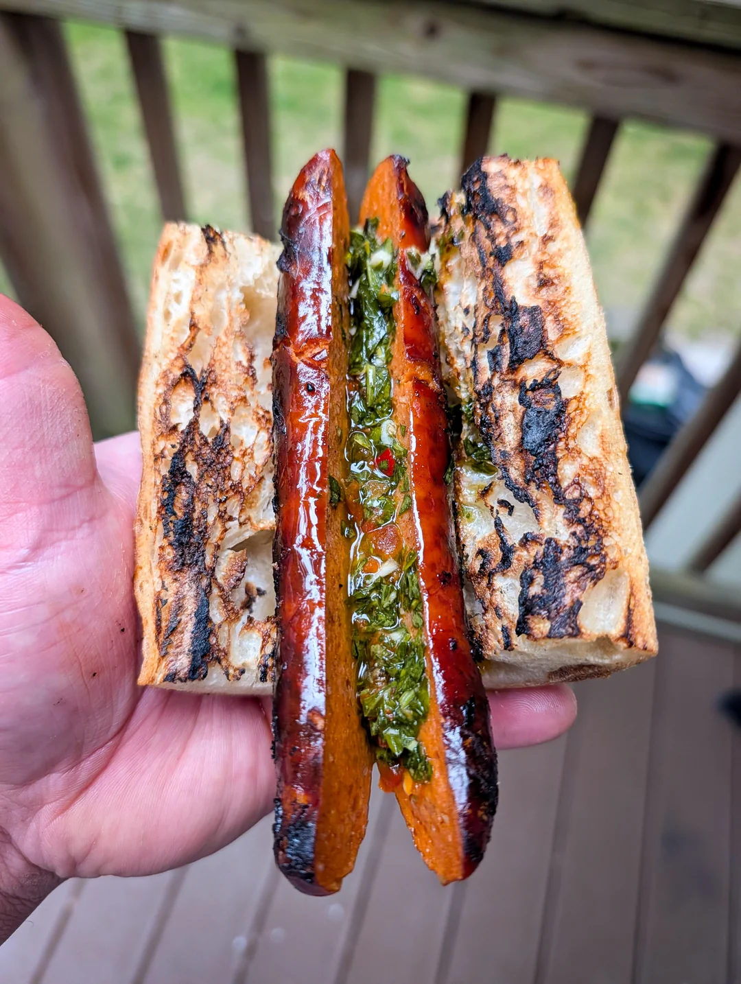 A hand holds a grilled sausage sliced in half on toasted bread, topped with green chimichurri sauce; wooden deck and railing are blurred in the background.