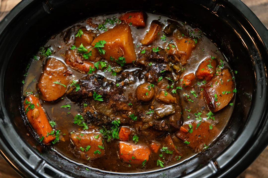 A slow cooker filled with beef stew, including chunks of beef, carrots, potatoes, and herbs in a rich brown broth, garnished with chopped parsley.