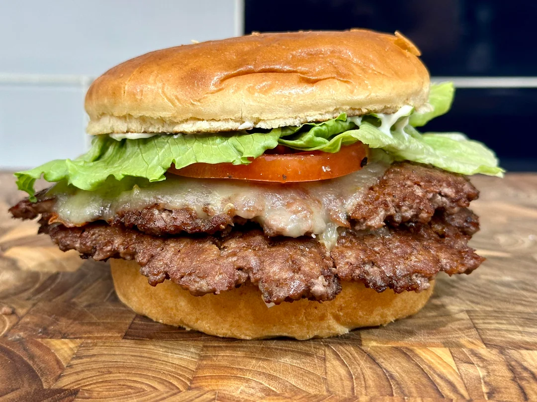 A cheeseburger with two beef patties, melted cheese, lettuce, tomato, and mayonnaise in a toasted bun, on a wooden surface.