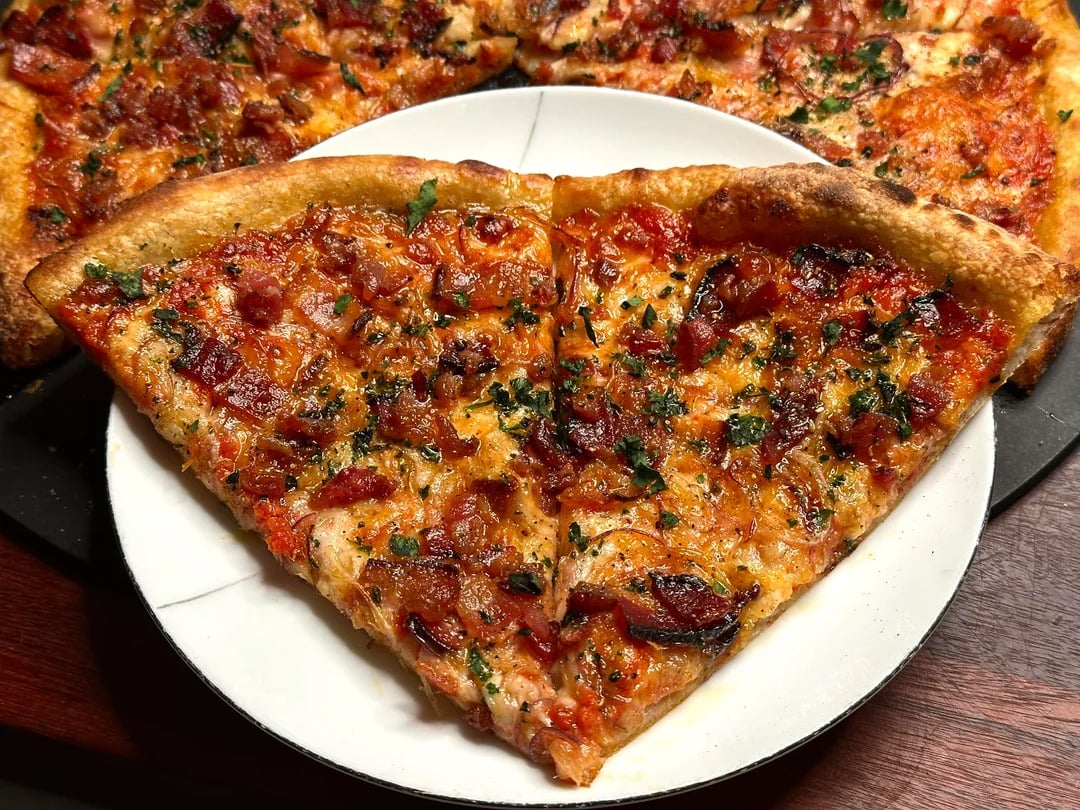 A close-up of a large pizza slice on a white plate, topped with melted cheese, bits of bacon, herbs, and a golden, crispy crust. More pizza is visible in the background.