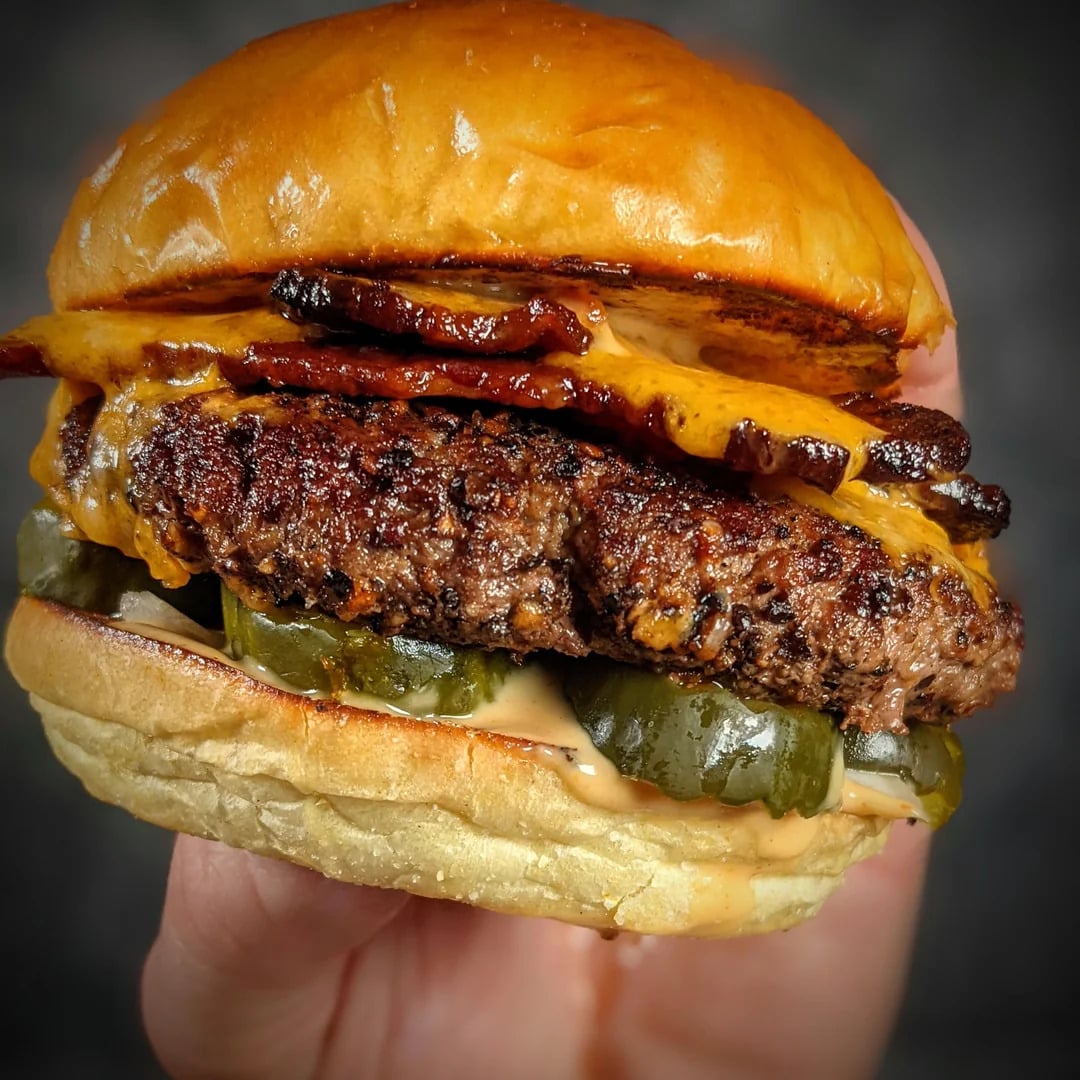 A hand holds a cheeseburger with a beef patty, cheddar cheese, bacon, pickles, and sauce in a shiny brioche bun.