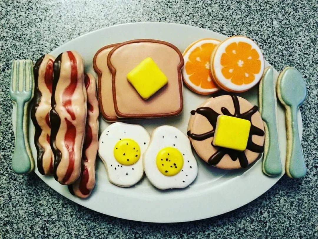 Cookies decorated as breakfast foods—bacon, eggs, toast, pancakes with butter, orange slices, and utensils—arranged on a white oval plate against a speckled countertop background.