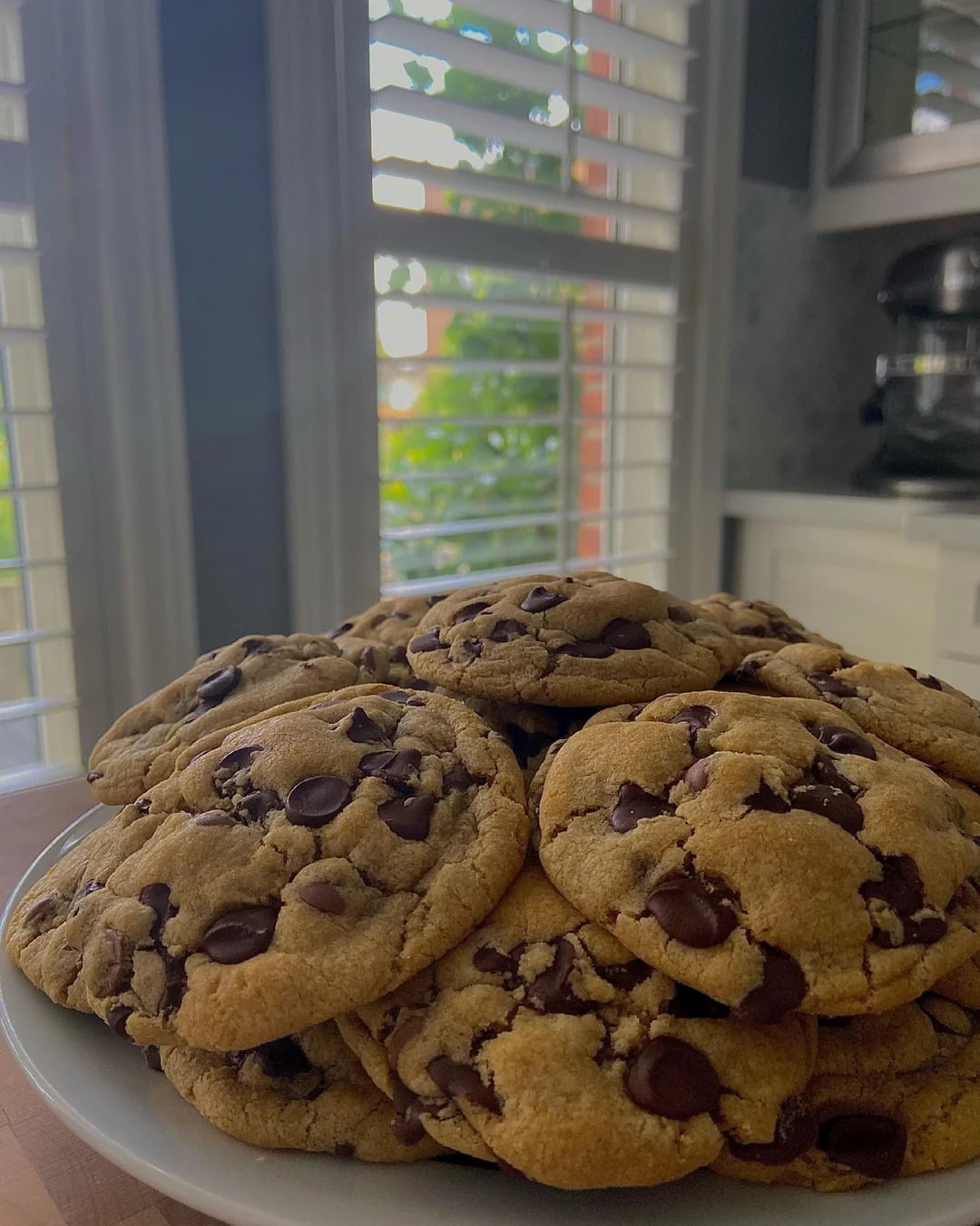 A plate piled high with chocolate chip cookies sits on a kitchen counter, with sunlight streaming through window blinds in the background.