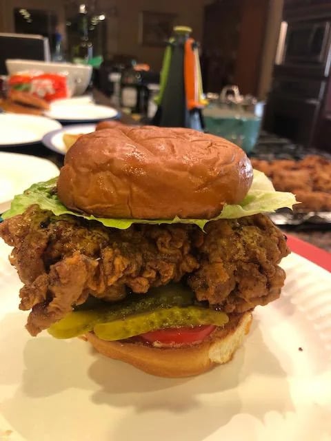 A close-up of a fried chicken sandwich on a plate, topped with lettuce, pickles, tomato, and a toasted bun, with a blurred kitchen background.