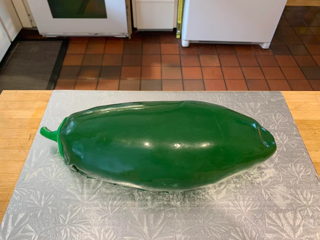 A realistic cake shaped and decorated to look like a large green jalapeño pepper sits on a patterned silver board in a kitchen.