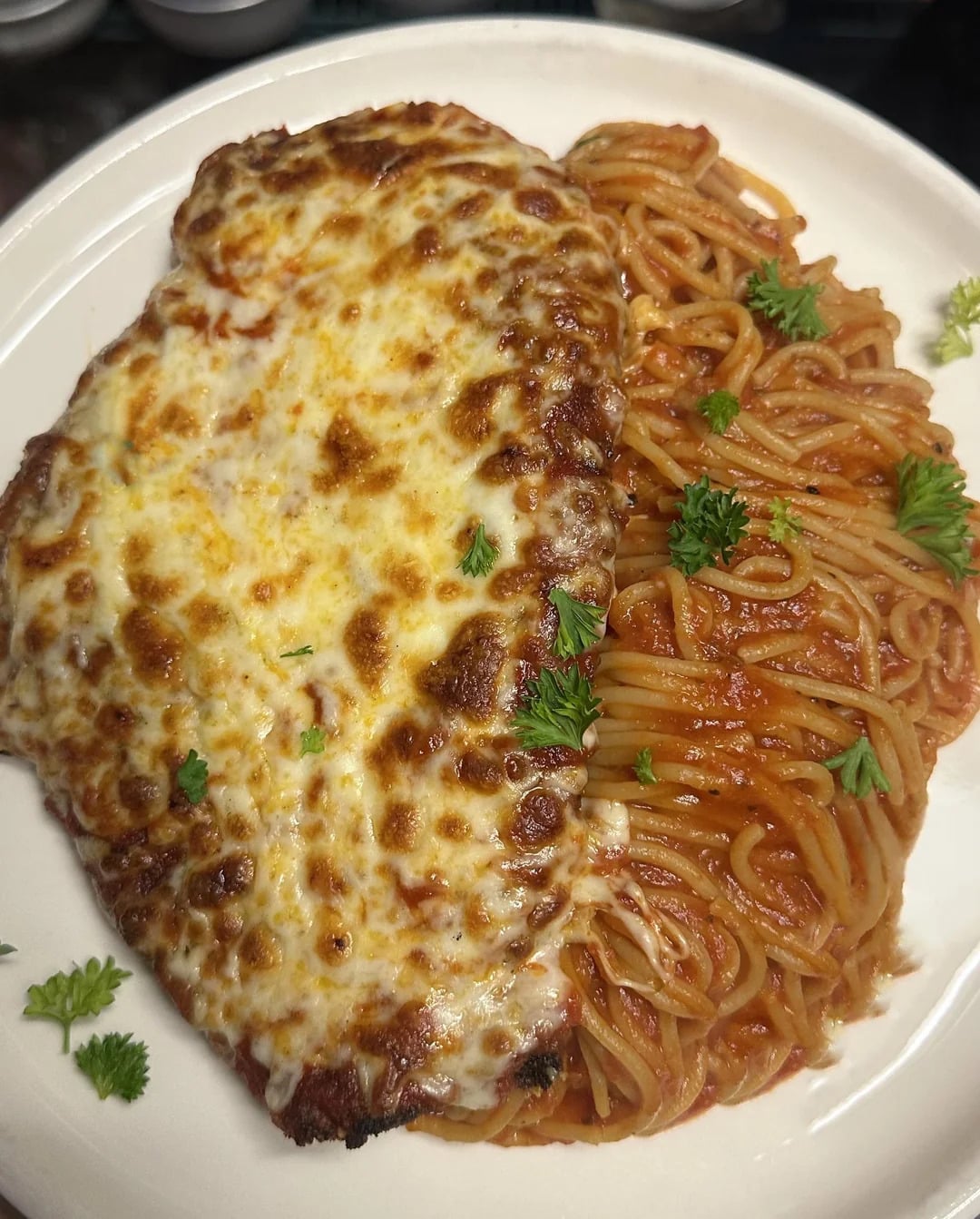 A plate with cheesy chicken Parmesan next to a portion of spaghetti in tomato sauce, both garnished with fresh parsley.
