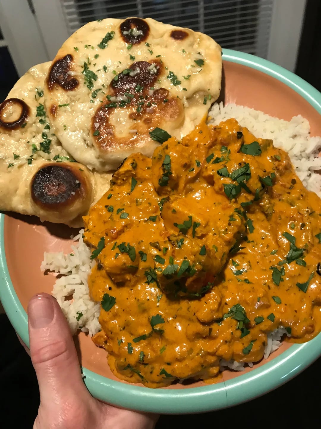 A hand holds a bowl with naan bread, white rice, and creamy orange chicken curry garnished with chopped herbs.