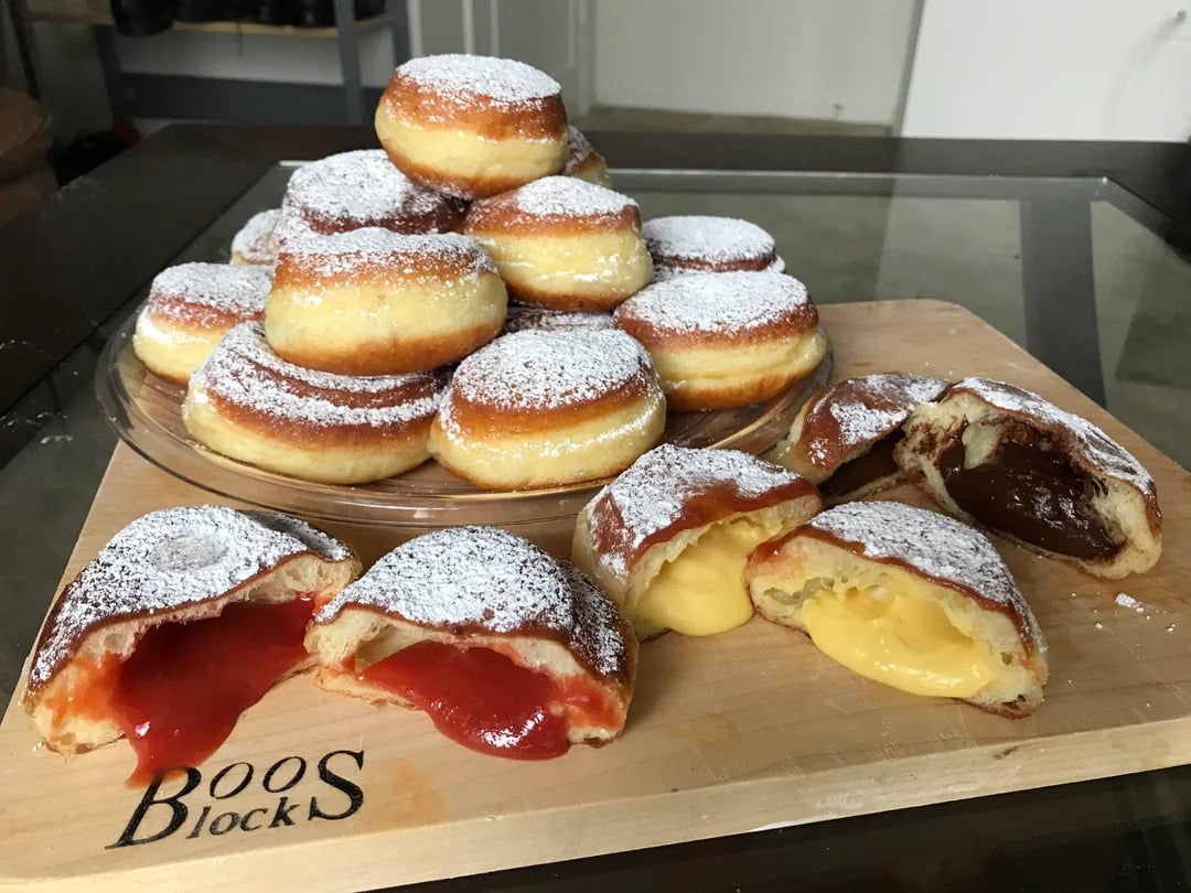 A plate of powdered jelly-filled and custard-filled donuts, with a few cut open to show red jelly, yellow custard, and chocolate fillings, sitting on a wooden cutting board labeled “Boos Blocks.”