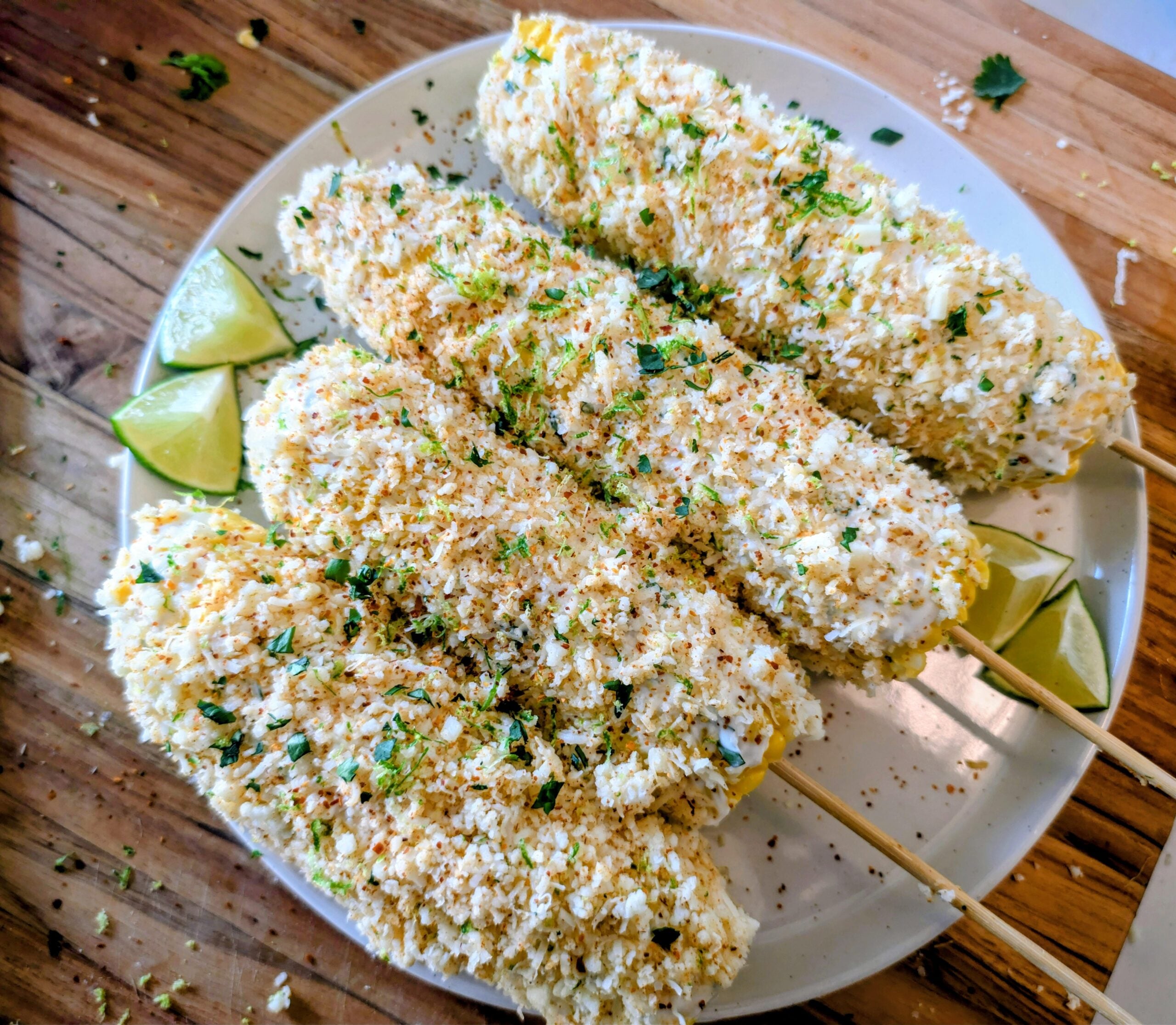 Four elotes (Mexican street corn) on skewers, coated with cheese and herbs, served on a white plate with lime wedges, on a wooden surface.