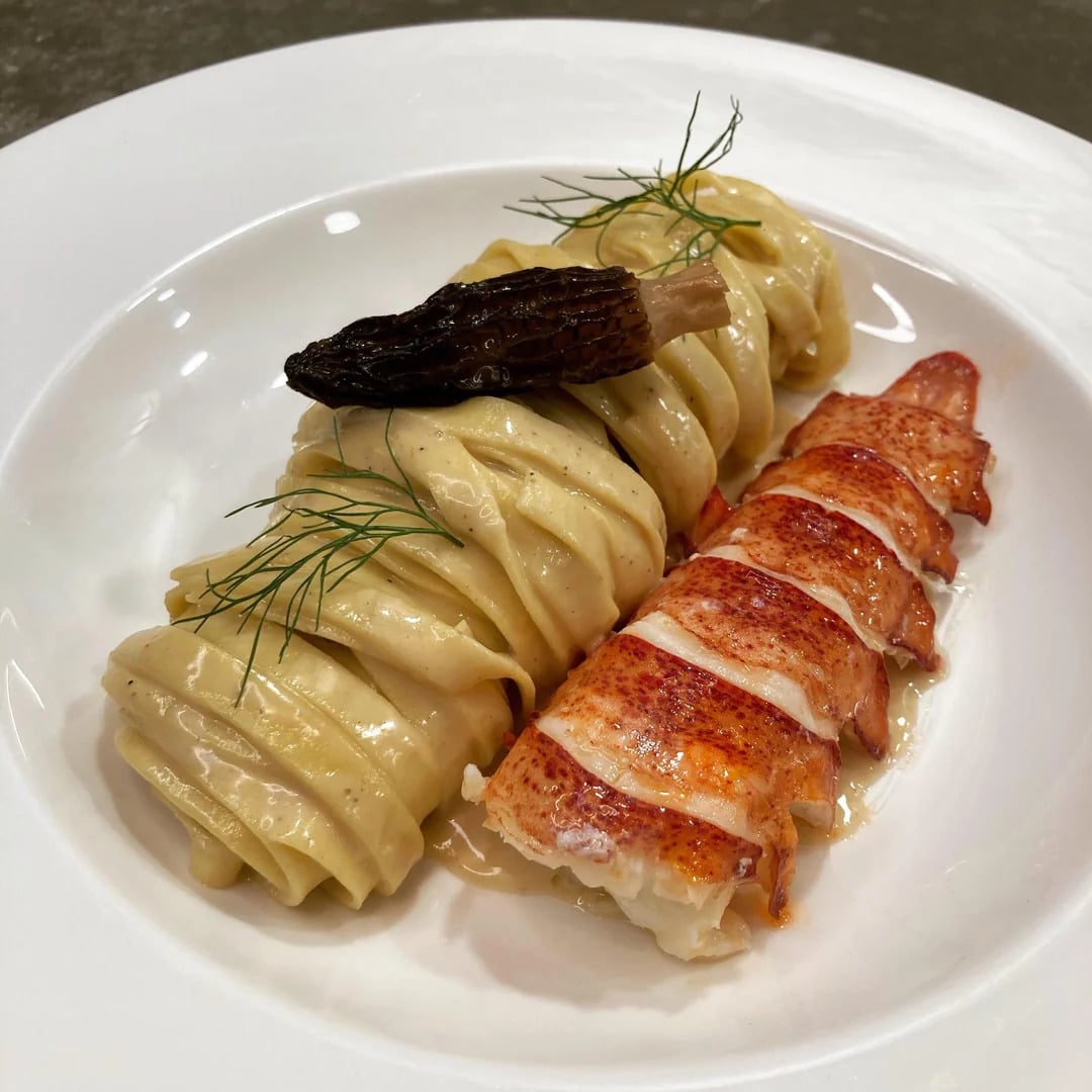Tagliatelle pasta topped with a morel mushroom and dill, served beside sliced lobster tail on a white plate.