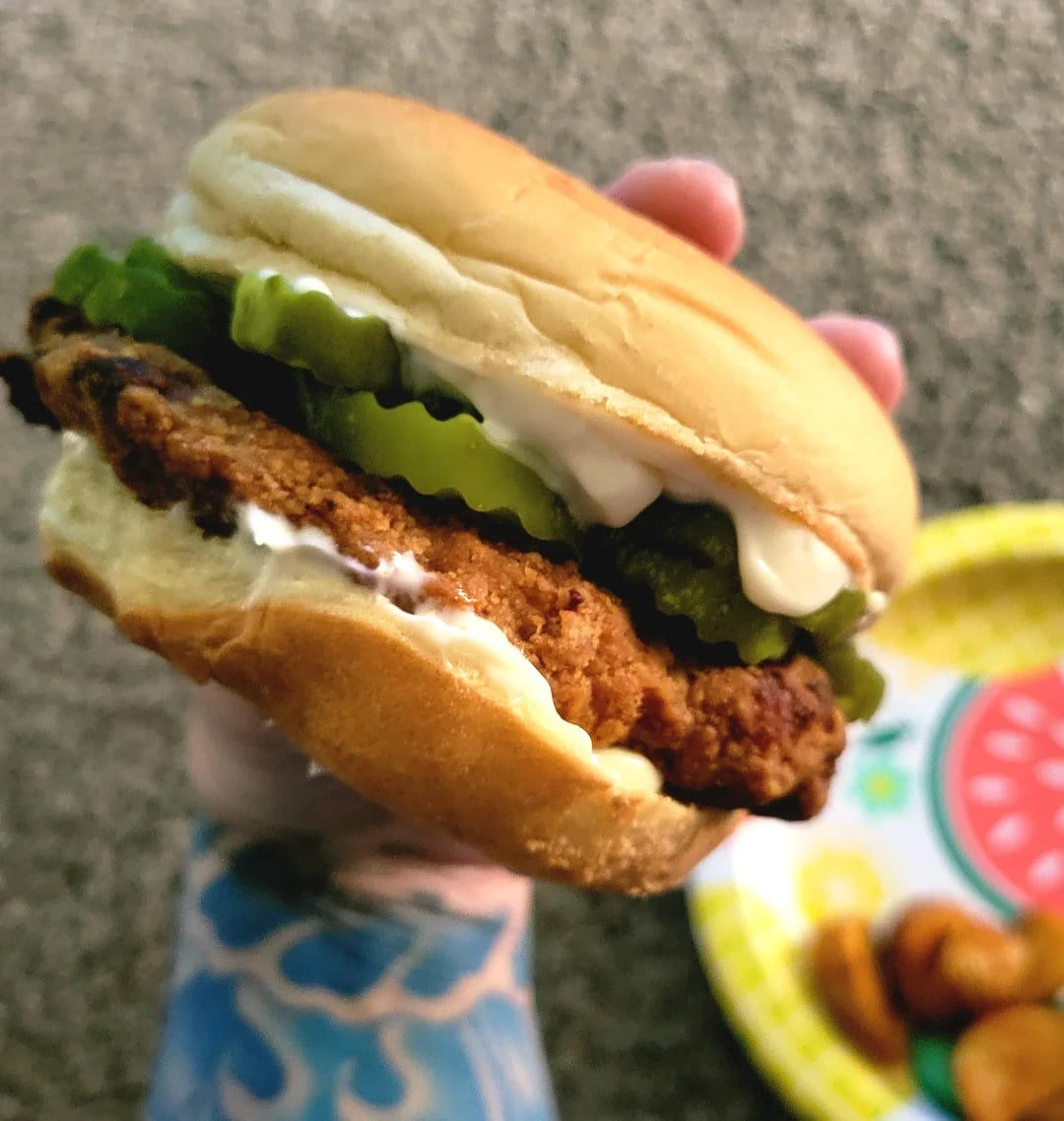 A hand holds a chicken sandwich with pickles and mayonnaise in a bun. In the background, a colorful plate with food sits on a textured surface.