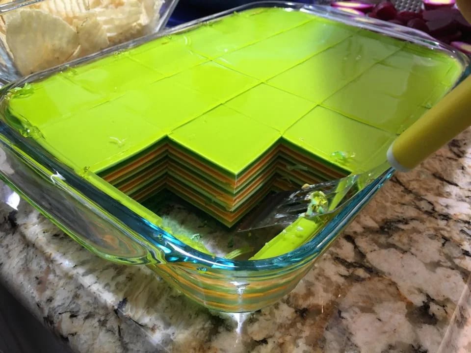 A glass dish holds neatly cut, multi-layered green and yellow gelatin squares, with a section removed to reveal the colorful layers inside. A serving utensil rests in the empty space.