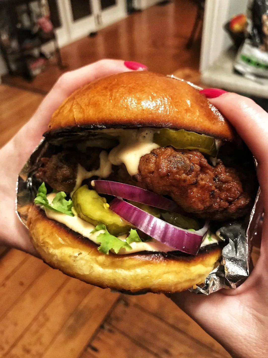 A close-up of hands holding a fried chicken sandwich with lettuce, pickles, red onion, and sauce in a shiny bun. The background shows a wooden floor and blurred kitchen items.