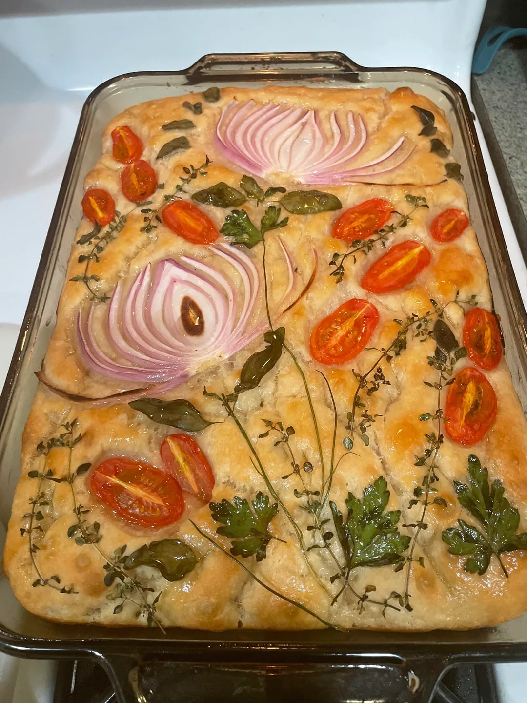 A rectangular focaccia bread decorated with sliced red onions, halved cherry tomatoes, and fresh herbs arranged to resemble flowers and stems.