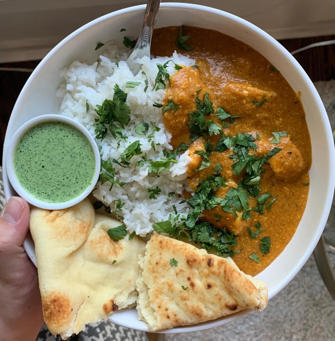 A bowl of white rice, orange curry, and cilantro, with naan bread and green sauce on the side. A fork is in the bowl.
