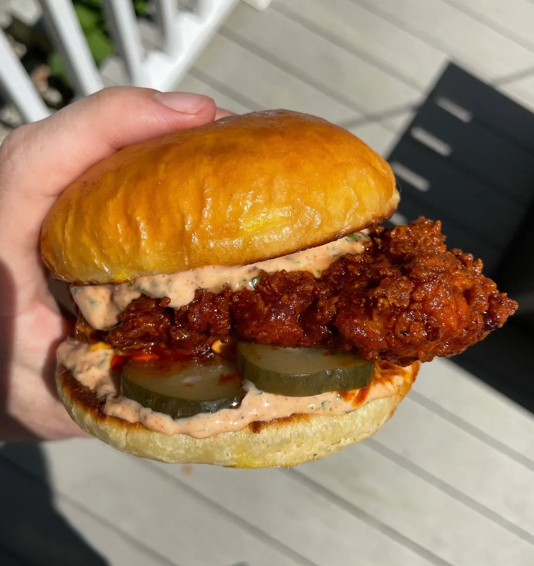A hand holds a fried chicken sandwich with pickles and creamy sauce in a shiny bun, shown close up in bright sunlight.
