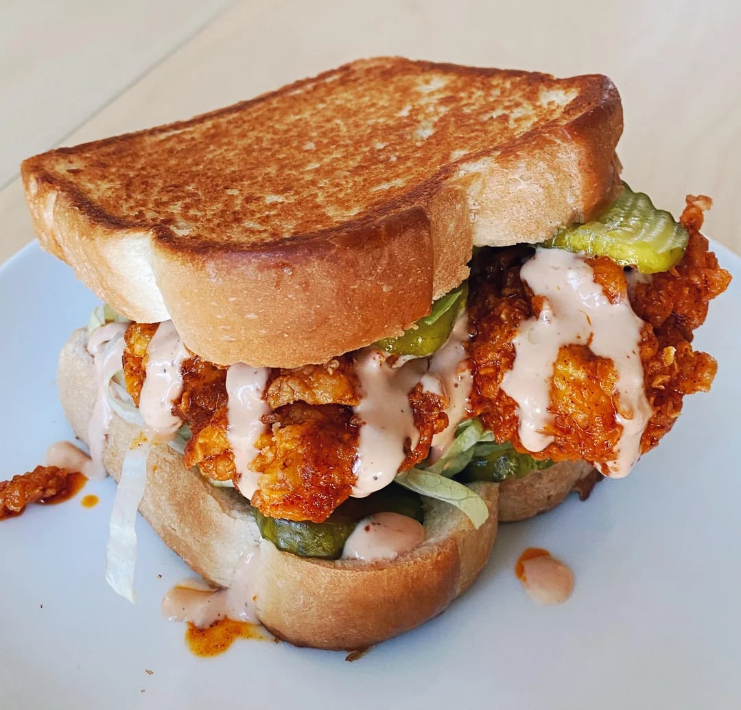 A fried chicken sandwich with pickles, shredded lettuce, and creamy sauce, served on toasted bread, sits on a white plate.