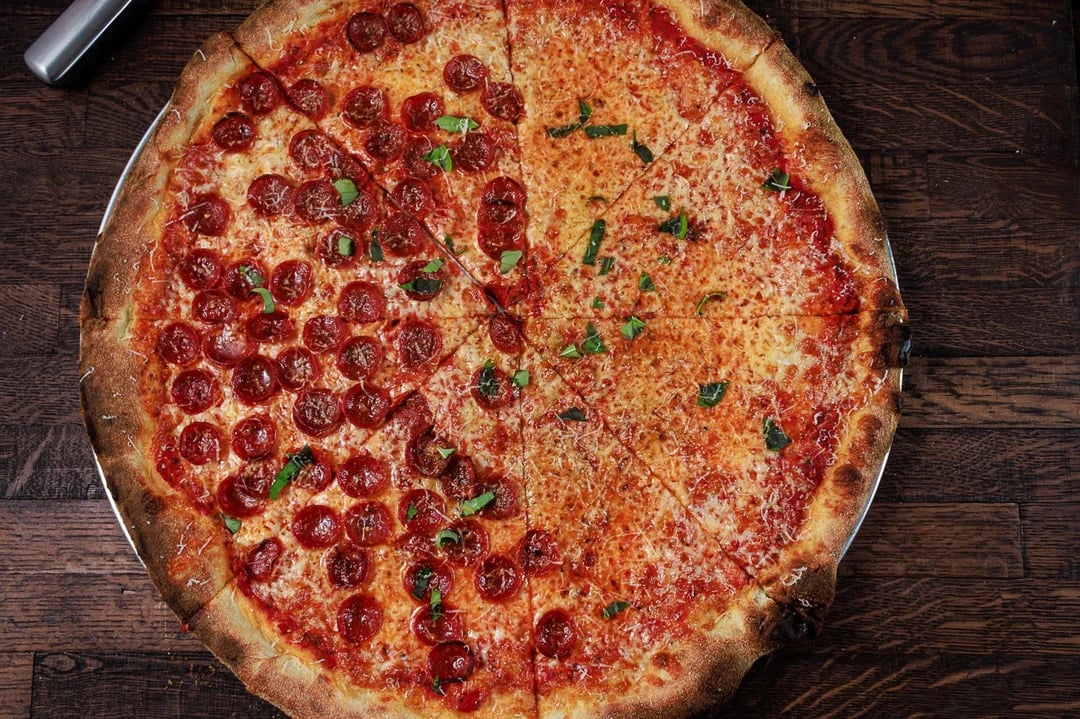 A large pizza on a wooden table, half topped with pepperoni slices and fresh basil, and the other half plain cheese with basil sprinkled on top. The pizza is cut into slices and served on a metal tray.