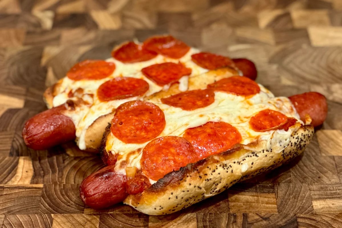 Two hot dogs in poppy seed buns, topped with melted cheese and pepperoni slices, placed on a wooden surface.