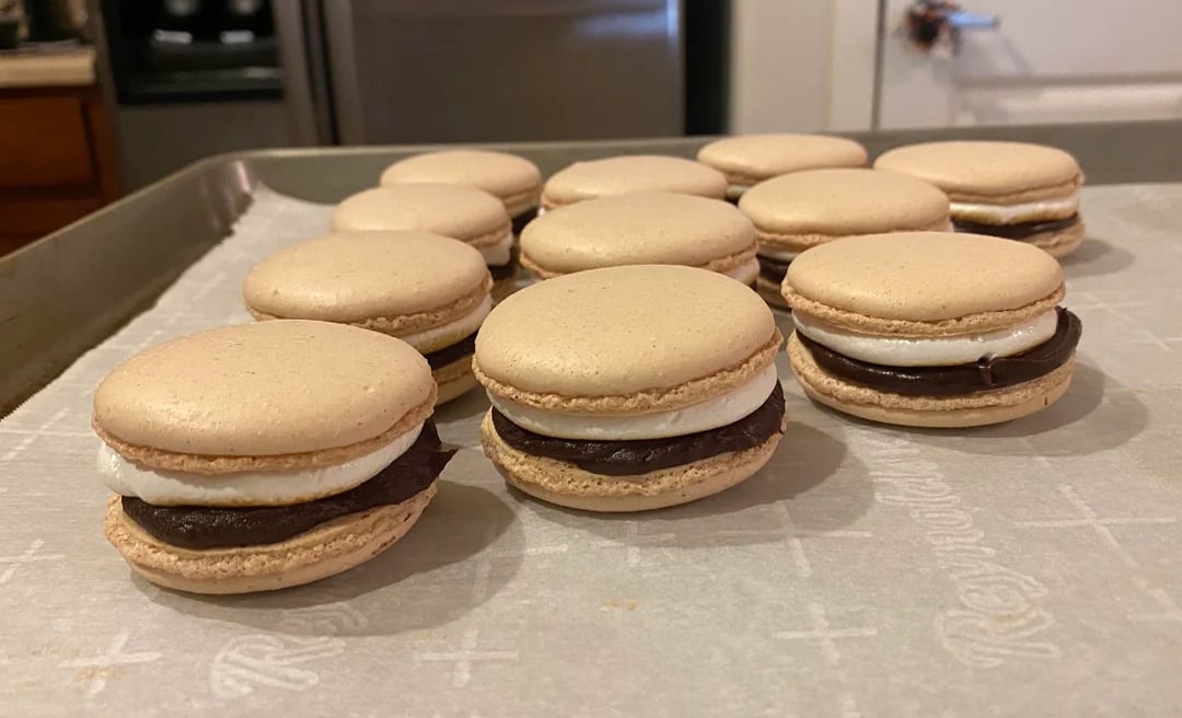 A tray of homemade s'mores macarons with chocolate and marshmallow filling sits on parchment paper in a kitchen.