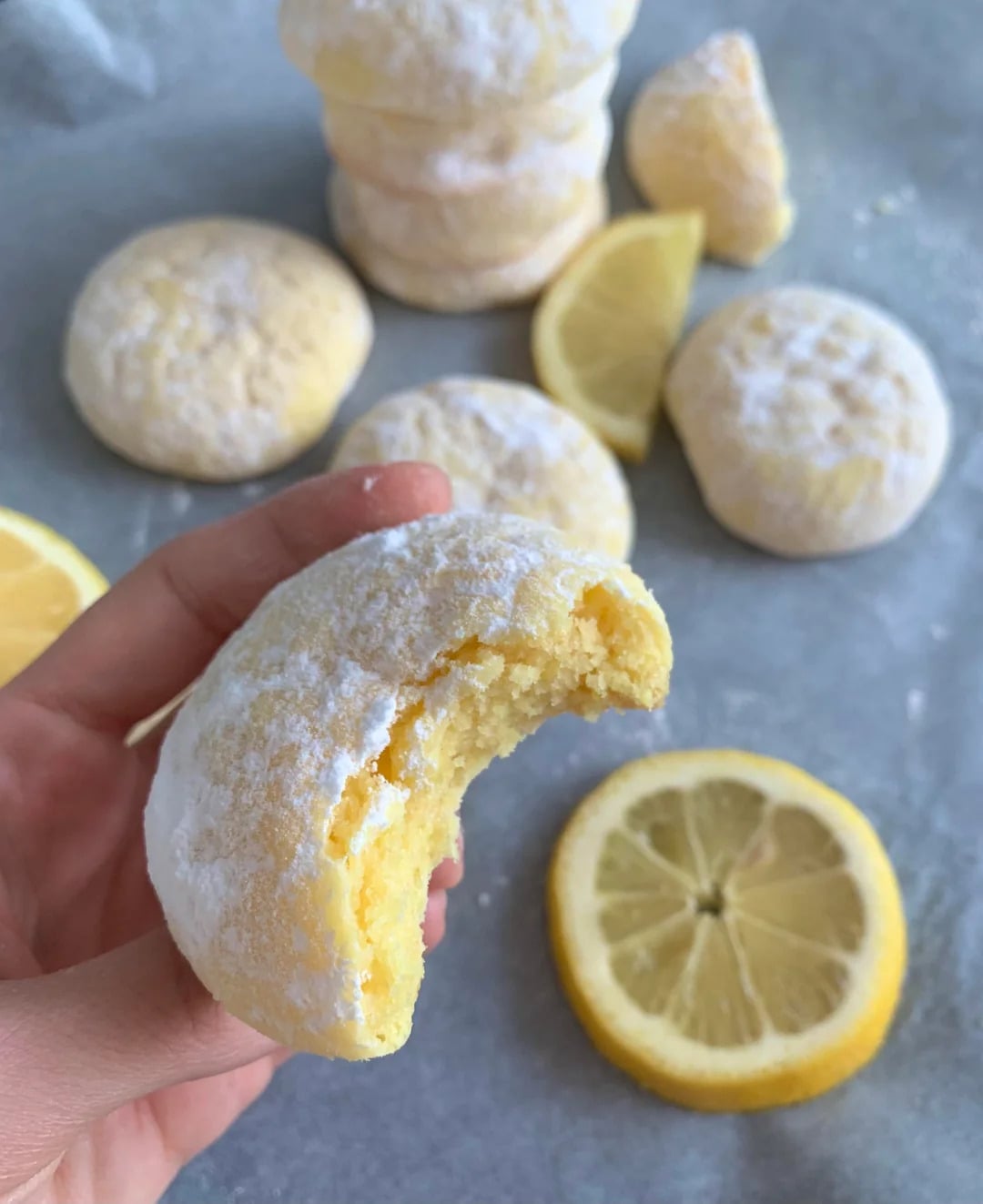 A hand holds a lemon cookie dusted with powdered sugar and a bite taken out. Other cookies and lemon slices are on a gray surface in the background.