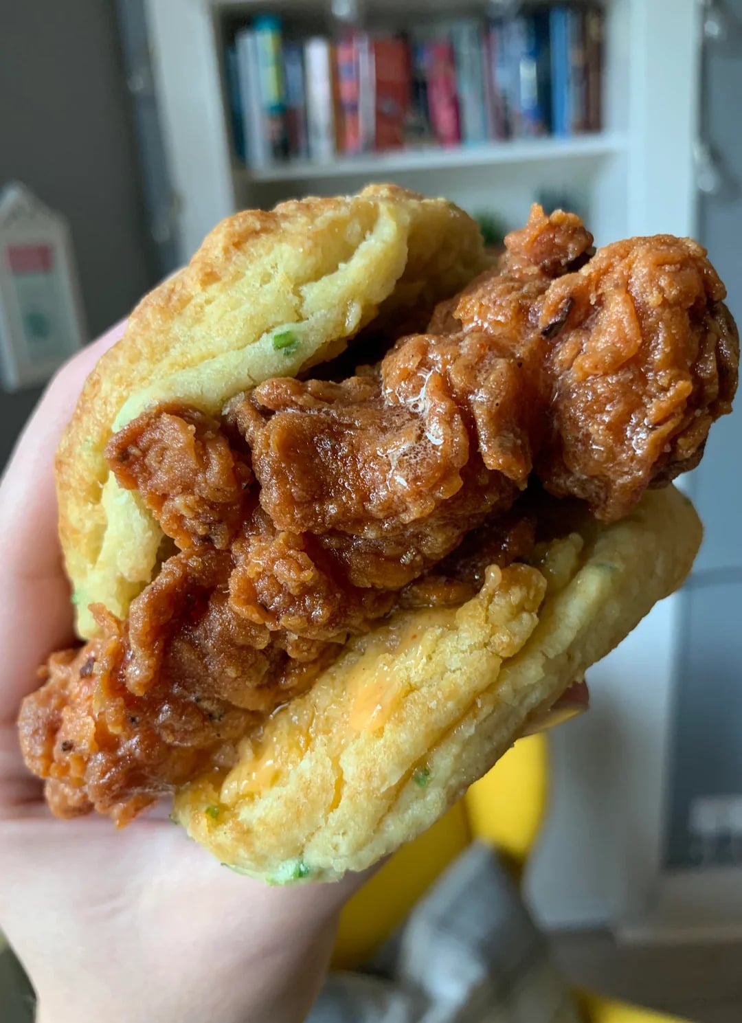 A hand holds a fried chicken biscuit sandwich with a crispy, golden-brown chicken fillet between fluffy, homemade biscuits. A bookshelf and room interior are blurred in the background.
