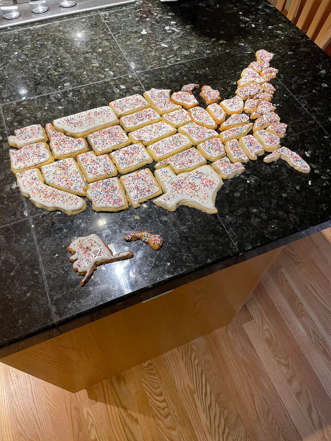 Cookies shaped and arranged like a map of the United States, each state decorated with white icing and colorful sprinkles, set on a dark countertop.